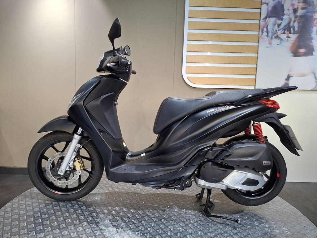 Piaggio Medley (2022) 17 A black scooter displayed in a showroom, featuring a sleek design with a visible seat, handlebars, and wheels, resting on a textured surface.