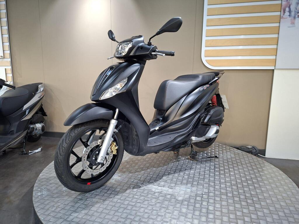Piaggio Medley (2022) 19 A black scooter is displayed on a circular platform in a modern showroom, with a minimalist background featuring wooden paneling. The scooter has a sleek design with a prominent front headlight and silver accents on the front wheel and brakes.