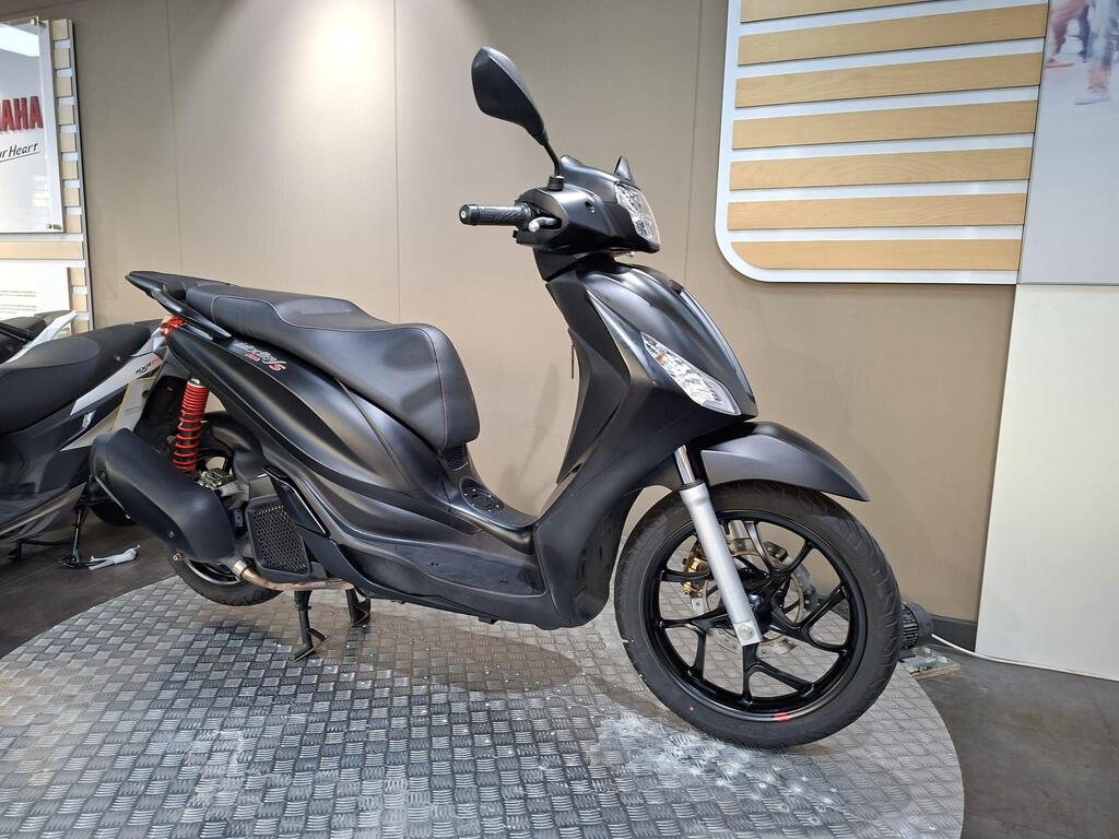 Piaggio Medley (2022) 2 A sleek black scooter displayed on a textured circular platform in a modern showroom, featuring a sporty design and red shock absorbers.