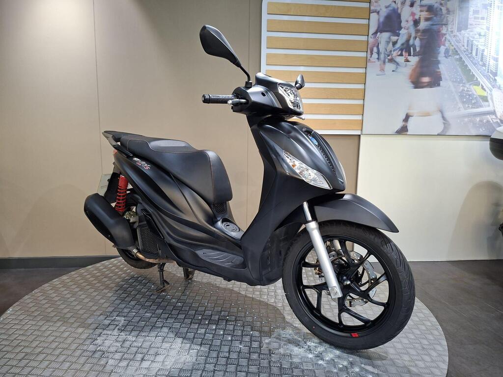 Piaggio Medley (2022) 24 A sleek black scooter displayed on a circular metallic platform in a minimalist showroom, featuring a prominent front headlight and detailed wheel design. The background includes a subtle design element, enhancing the modern aesthetic.