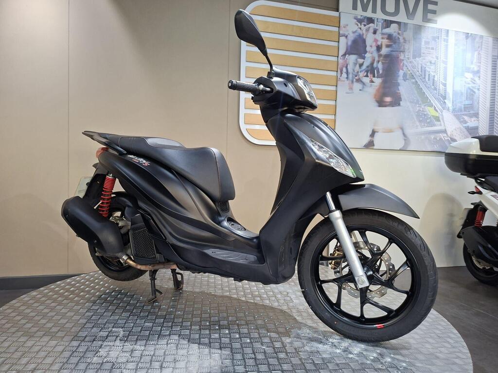 Piaggio Medley (2022) 26 A sleek black scooter is displayed on a circular platform, showcasing its design features like red suspension springs and black wheels, set against a modern background with a cityscape.