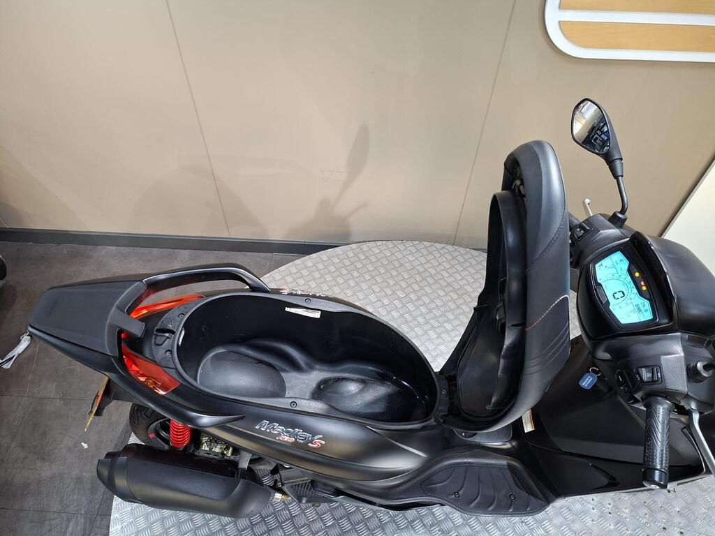 Piaggio Medley (2022) 6 A black scooter sits on a gray textured platform, showcasing its seat and dashboard. The dashboard features a blue digital display with various indicators, while the scooter’s storage compartment is open, revealing its interior. The background is a neutral-colored wall.