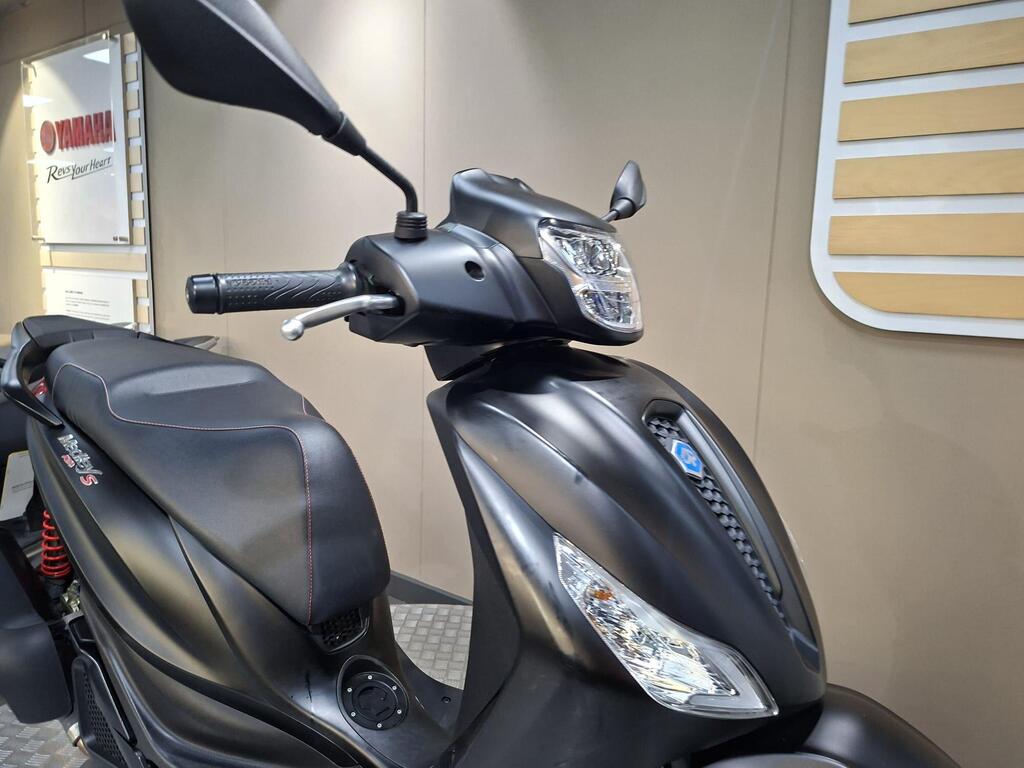 Piaggio Medley (2022) 8 A close-up view of a sleek black scooter, highlighting the handlebars, mirror, and part of the seat. The scooter features modern design elements and is displayed against a neutral background with branding visible.