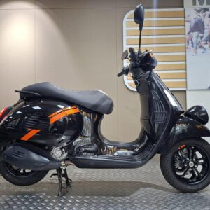 Vespa GTV 300 (2025) A black scooter with orange accents is displayed on a raised platform against a neutral background. The scooter features a rounded body, a comfortable seat, and sleek wheels.