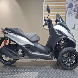 Piaggio MP3 300 HPE Sport (2022) A three-wheeled scooter in silver and black, featuring a prominent windshield, sleek design, and a red suspension system, displayed on a circular platform against a neutral wall background.
