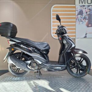 SYM Symphony ST 125 (2015) A sleek black scooter with a rear trunk stands on a textured circular platform, set against a neutral background with a partially visible urban scene.