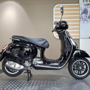 Vespa GTS Super (2023) A black scooter sits on a circular, textured platform against a simple background. The scooter features a shiny chrome exhaust, a smooth seat, and is viewed from the side, showcasing its sleek design and modern wheels.