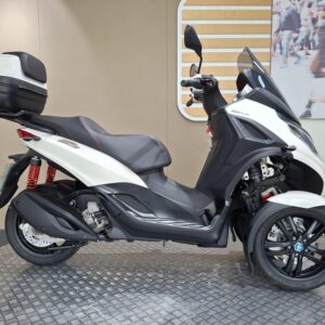 Piaggio MP3 300 Sport (2021) A white three-wheeled scooter with a black seat and a top case, positioned on a textured surface against a neutral background with abstract designs.