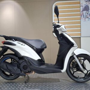 Piaggio Liberty S 125 (2025) A side view of a white scooter with black accents, featuring a sleek design, visible wheels, and a modern seat, displayed on a textured surface against a neutral background.