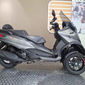 Piaggio MP3 350 LT Sport (2019) A side view of a gray three-wheeled scooter displayed on a textured floor, with a neutral background featuring a branding element and blurred people.