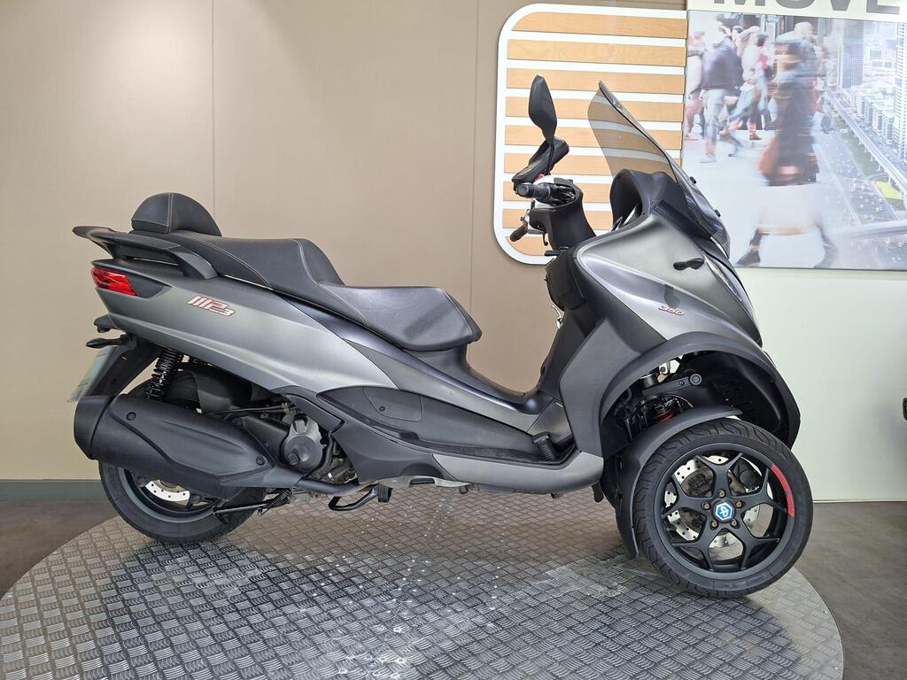 Piaggio MP3 350 2019 1 Piaggio MP3 350 LT Sport (2019) A side view of a gray three-wheeled scooter displayed on a textured floor, with a neutral background featuring a branding element and blurred people.