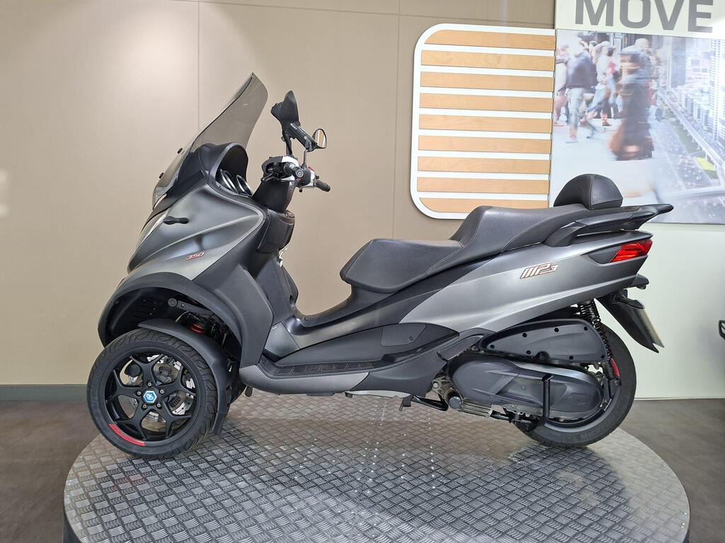 Piaggio MP3 350 2019 10 A sleek gray three-wheeled scooter showcased on a circular black platform in a modern display setting. The scooter features a visor, comfortable seating, and sporty wheels, with a background partially visible showing a bustling street scene.