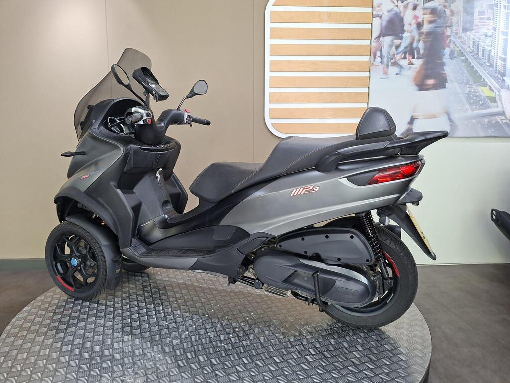 Piaggio MP3 350 2019 11 A sleek, three-wheeled scooter displayed in a showcase, featuring a dark gray body, large windshield, and prominent rear lights. The background includes a neutral-colored wall with a modern graphic of a city scene.