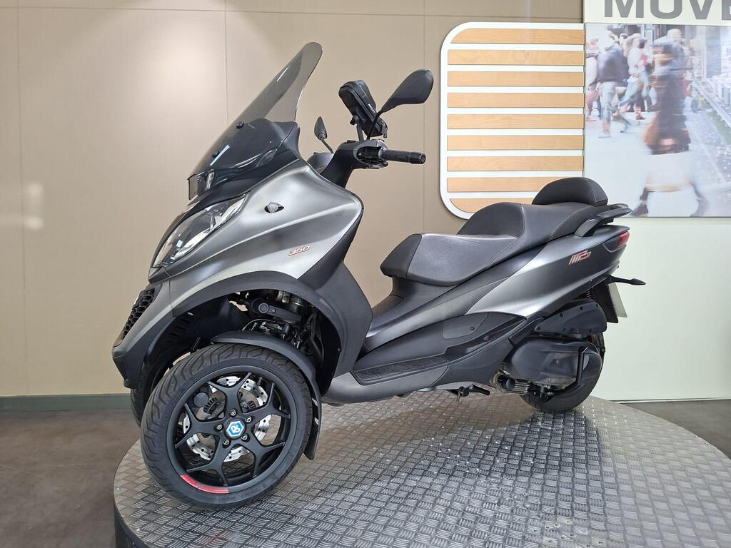 Piaggio MP3 350 2019 12 A grey three-wheeled scooter displayed on a circular platform with a textured surface, featuring a large windshield, sporty design, and a prominent logo on the front wheel. In the background, there is a blurred image of shoppers.