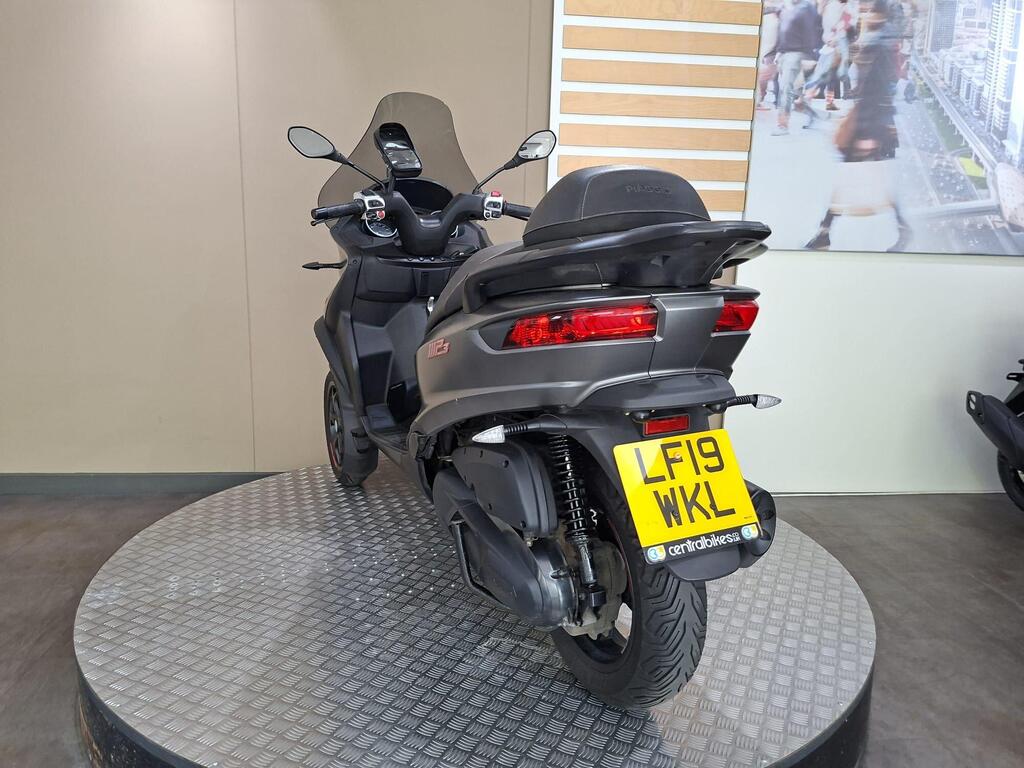 Piaggio MP3 350 2019 13 A gray three-wheeled scooter displayed on a rotating platform, featuring a rear view with a distinctive design, tail lights, and a license plate reading "LF19 WKL." The background shows a neutral wall with wooden elements.