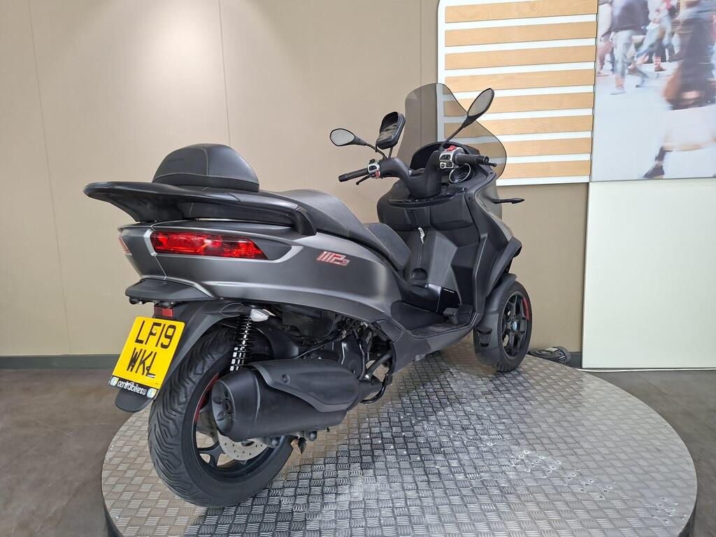 Piaggio MP3 350 2019 15 A sleek, dark gray three-wheeled scooter displayed on a circular platform, with seating for two and modern design elements, set against a neutral wall backdrop and a decorative panel.