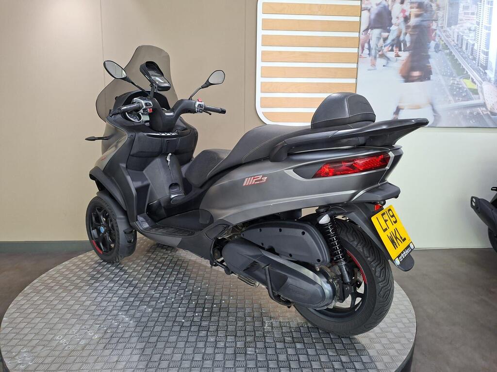 Piaggio MP3 350 2019 16 A grey three-wheeled scooter, model MP3 500, positioned on a circular platform with a patterned surface. The scooter features a rear view with distinctive red accents and a license plate displaying "LF19 WKL." The background shows a neutral-colored wall with a blurred city scene.