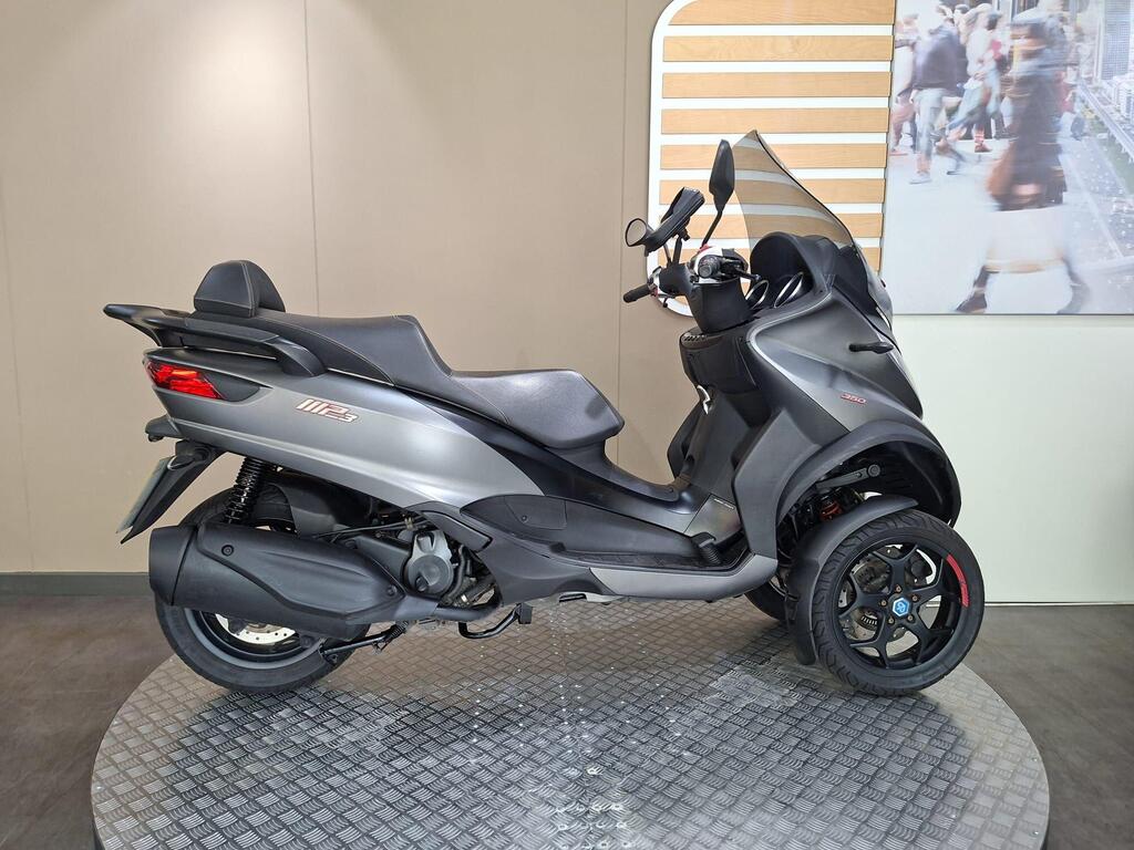 Piaggio MP3 350 2019 17 A side view of a gray three-wheeled scooter displayed on a circular platform with a simple light-colored background.