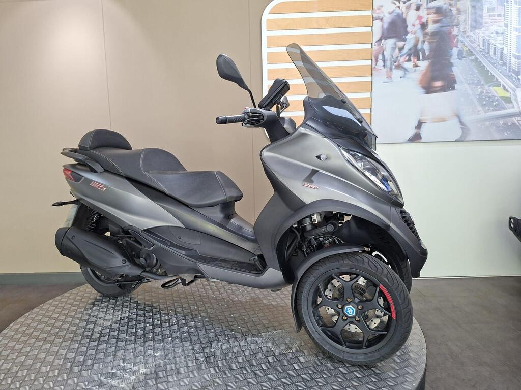 Piaggio MP3 350 2019 2 A gray three-wheeled scooter with stylish design features, including a tinted windshield, is displayed on a textured circular platform in front of a minimalist backdrop.