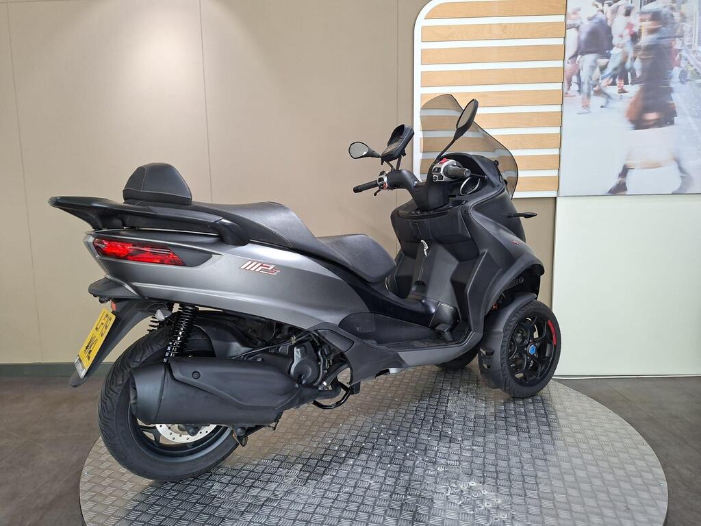 Piaggio MP3 350 2019 20 A gray three-wheeled scooter displayed on a circular platform, with a modern showroom background featuring a large image of people walking. The scooter has a sleek design, prominent rear lights, and a small windshield.