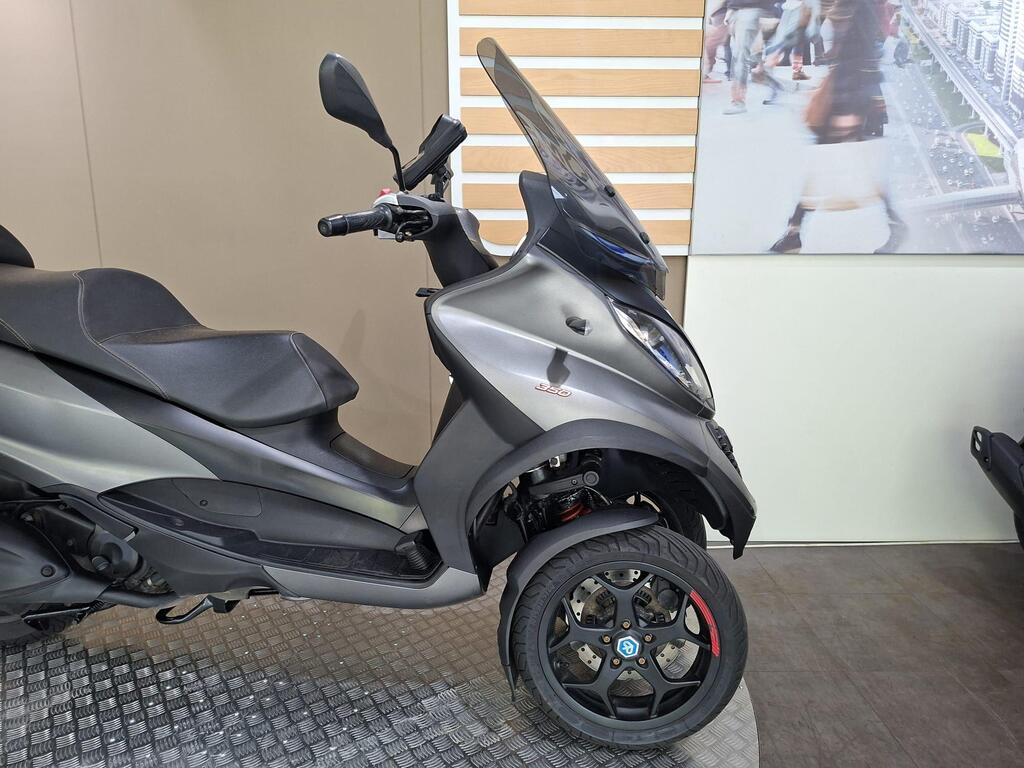Piaggio MP3 350 2019 22 A parked gray scooter with a sleek design, featuring two front wheels and a black seat, positioned on a textured floor in a showroom.