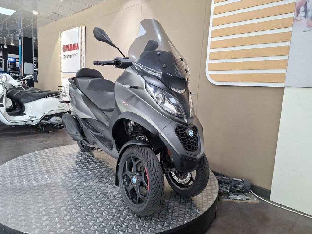 Piaggio MP3 350 2019 23 A three-wheeled scooter with a sleek gray design is displayed on a circular stand in a showroom, with a Yamaha sign visible in the background.
