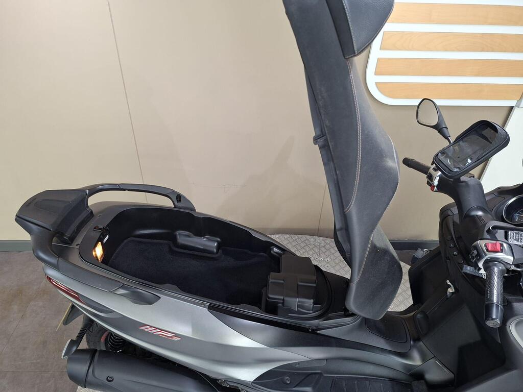 Piaggio MP3 350 2019 24 Close-up view of a motorcycle scooter's seat in an upright position, revealing a storage compartment underneath, with a black interior and a dashboard featuring controls and a digital display.