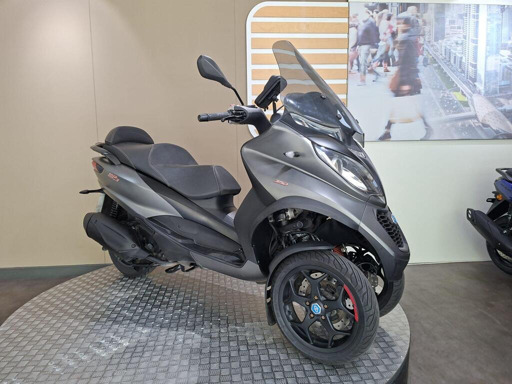 Piaggio MP3 350 2019 3 A sleek gray three-wheeled scooter displayed on a circular metal platform, featuring a protective windshield and sport-style wheels, set against a light-colored wall with a blurred cityscape image in the background.