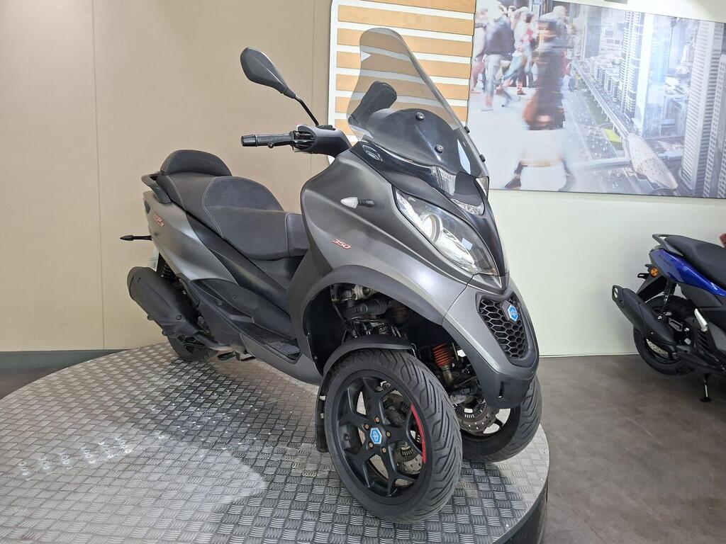 Piaggio MP3 350 2019 4 A dark gray three-wheeled scooter displayed on a circular platform in a showroom, with a modern interior and a blurred background featuring a city scene.