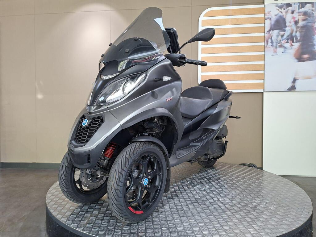 Piaggio MP3 350 2019 6 A gray three-wheeled scooter displayed on a circular platform, featuring a tinted windshield and distinct black alloy wheels. The background includes a blurred image of people walking.