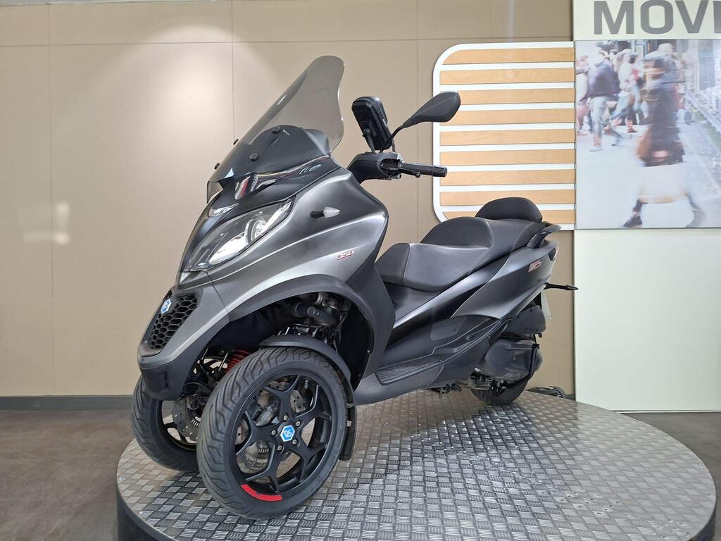 Piaggio MP3 350 2019 7 A black three-wheeled scooter displayed on a circular platform, featuring a windshield, large front headlights, and sporty wheels, with a beige wall and an action shot of people in the background.