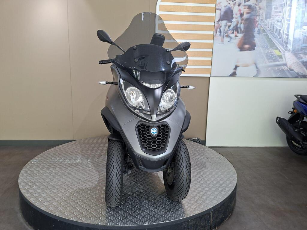 Piaggio MP3 350 2019 8 A gray three-wheeled scooter displayed on a circular platform, with a transparent windscreen and a modern design, against a neutral colored wall.