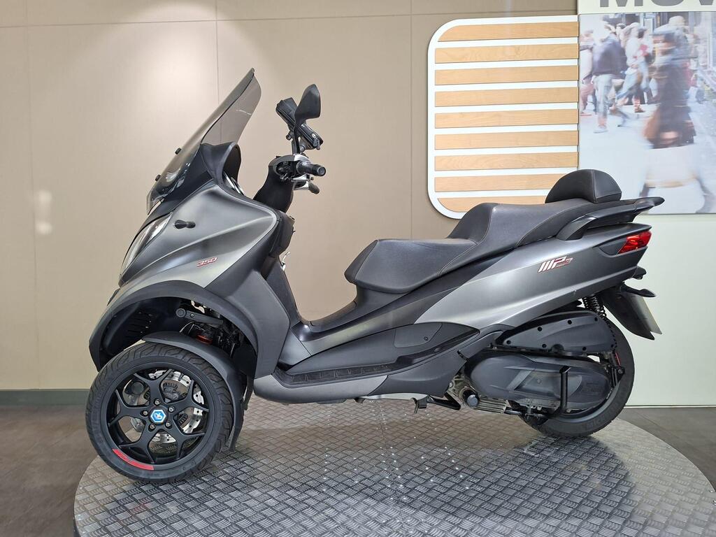 Piaggio MP3 350 2019 9 A sleek gray scooter with three wheels, featuring a tinted windshield and sporty black alloy wheels, displayed on a circular platform against a neutral background.