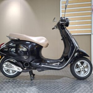 Vespa Primavera 50 (2017) A black scooter with a brown seat is positioned on a textured platform, set against a neutral background with a hint of people in motion visible in a nearby display.