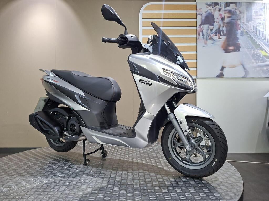 Aprilia SXR 50 2026 1 A silver Aprilia scooter displayed on a circular platform, with a neutral background featuring a blurred urban scene.