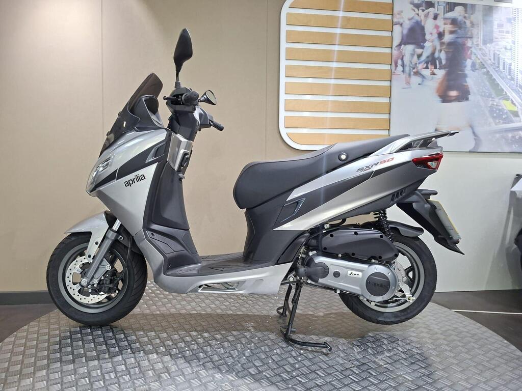 Aprilia SXR 50 2026 10 A silver Aprilia scooter parked on a textured platform against a neutral wall background. The scooter features a sleek design with a prominent windshield and a modern body style.