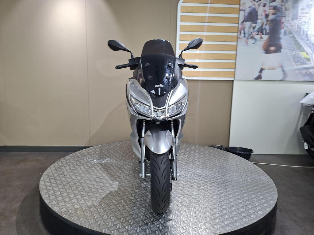 Aprilia SXR 50 2026 11 A silver motorcycle displayed on a rotating platform against a neutral background, featuring a modern design with a prominent front view, headlights, and mirrors.