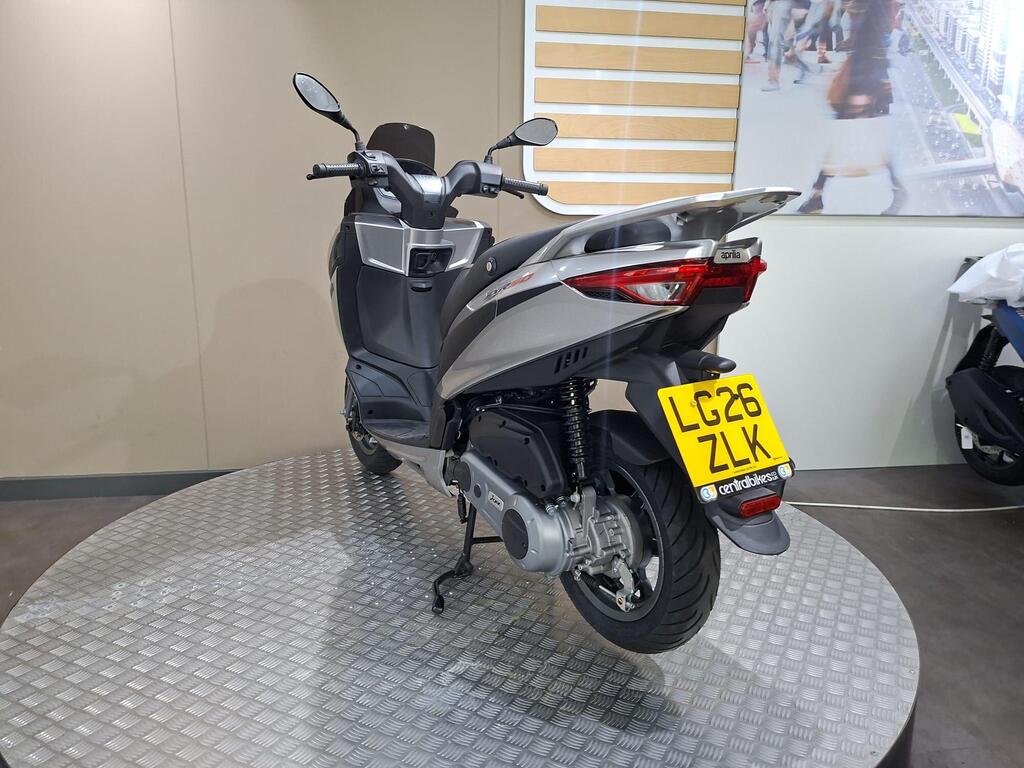 Aprilia SXR 50 2026 12 A silver scooter displayed in a showroom setting, featuring a clear view of its rear, side, and license plate. The scooter is set on a circular textured platform with a neutral background.