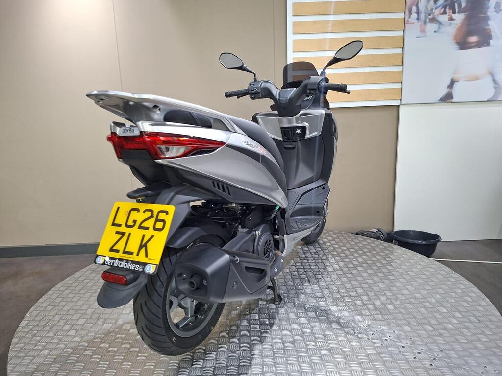 Aprilia SXR 50 2026 13 A silver Aprilia scooter positioned on a circular display platform with a textured surface, showcasing its rear view, including the license plate "LG26 ZLK." The background features a plain wall with horizontal stripes.