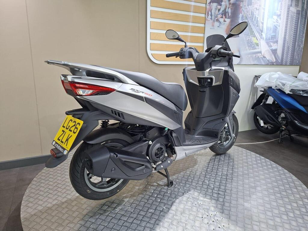 Aprilia SXR 50 2026 15 A modern silver and black scooter displayed on a circular metallic platform in a showroom, with a blurred background featuring a cityscape and another scooter partially visible. The scooter has a license plate visible.