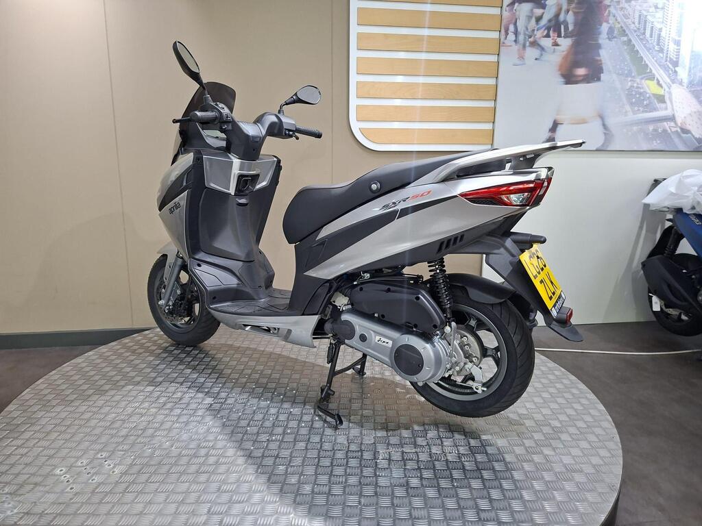 Aprilia SXR 50 2026 16 A sleek gray scooter displayed on a circular metal platform, with a backdrop of an indoor setting featuring modern design elements.