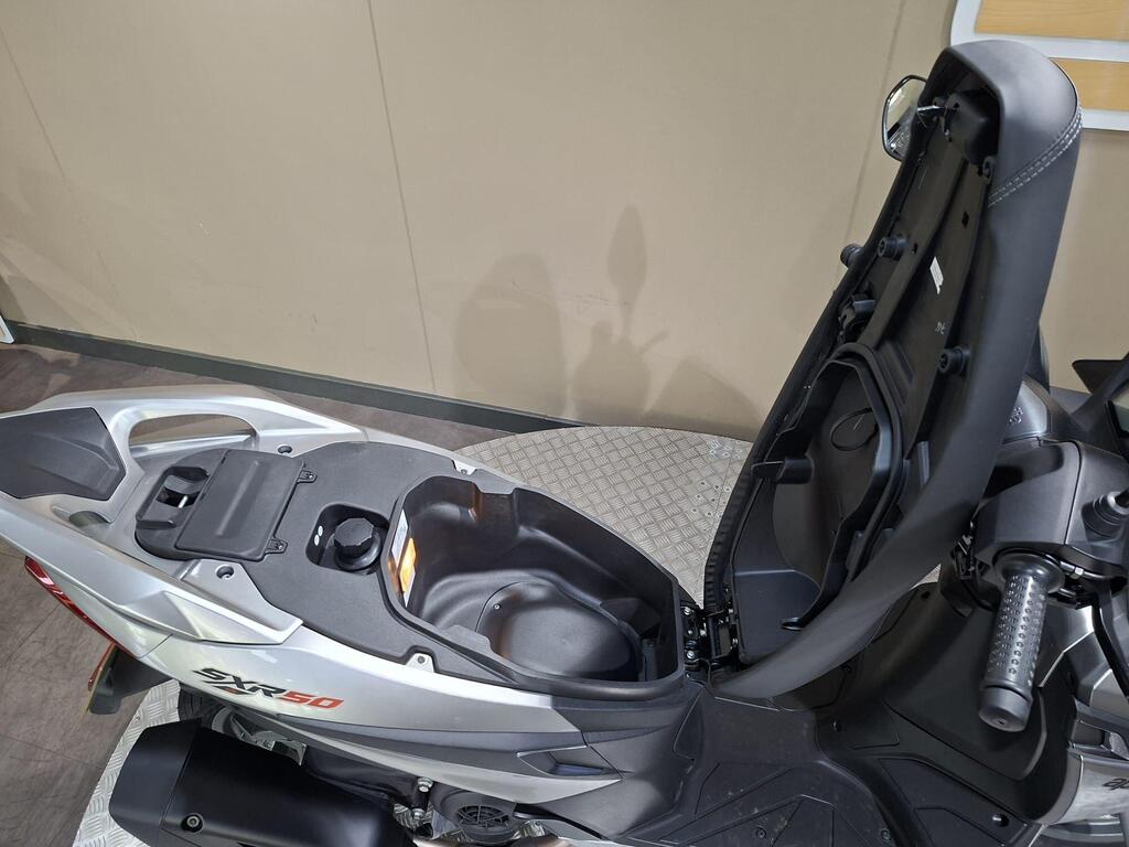 Aprilia SXR 50 2026 17 A close-up view of the seat compartment of an SRX 50 scooter, showing the area beneath the seat with storage options and a partially open lid. The background features a light-colored wall.