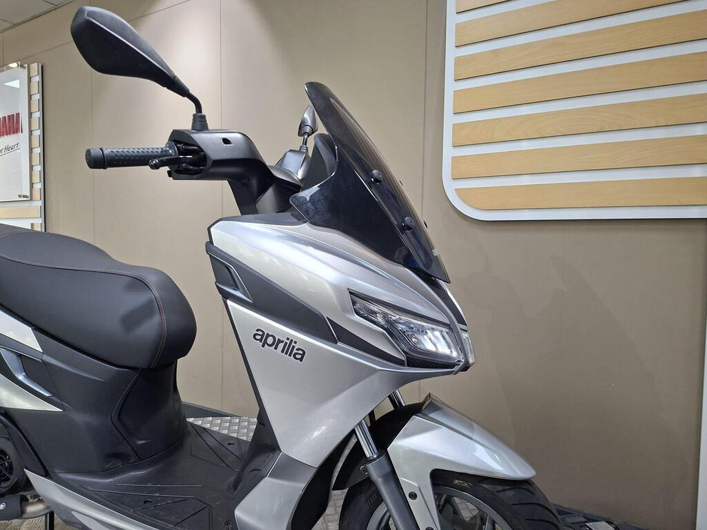 Aprilia SXR 50 2026 19 A close-up view of a silver Aprilia scooter, showcasing its sleek design, handlebar, and windshield, with a blurred background of a display wall.
