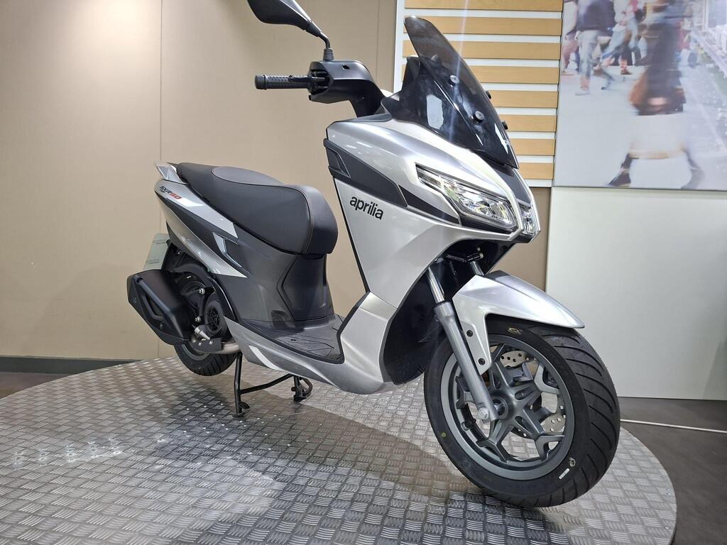 Aprilia SXR 50 2026 2 A silver Aprilia scooter displayed on a circular textured stand in a showroom setting. The scooter features a sleek design with a black seat and windscreen. A blurred background image of pedestrians is visible behind the scooter.