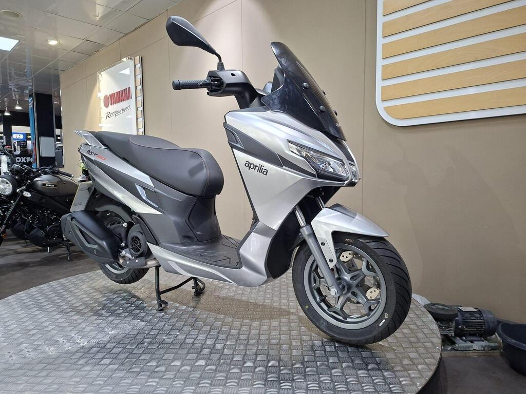 Aprilia SXR 50 2026 20 A silver Aprilia scooter is displayed on a patterned platform in a showroom, with a blurred black motorcycle visible in the background.