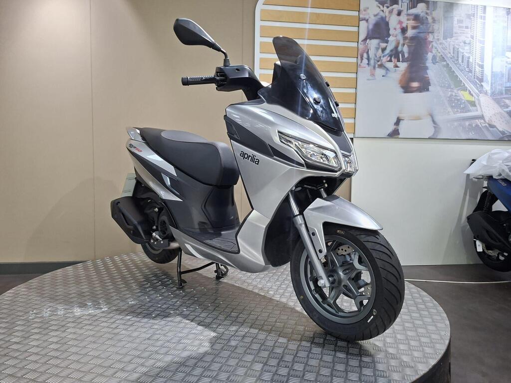 Aprilia SXR 50 2026 3 A silver Aprilia scooter displayed on a circular platform, featuring a tall windscreen and modern design elements, with a blurred backdrop of people and urban scenery.
