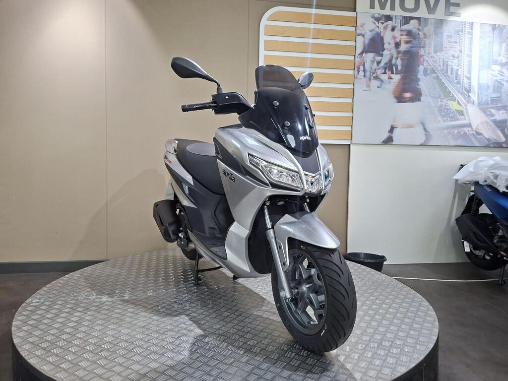 Aprilia SXR 50 2026 4 A sleek silver scooter is displayed on a circular platform, with a modern interior background featuring branding and a blurred image of pedestrians.