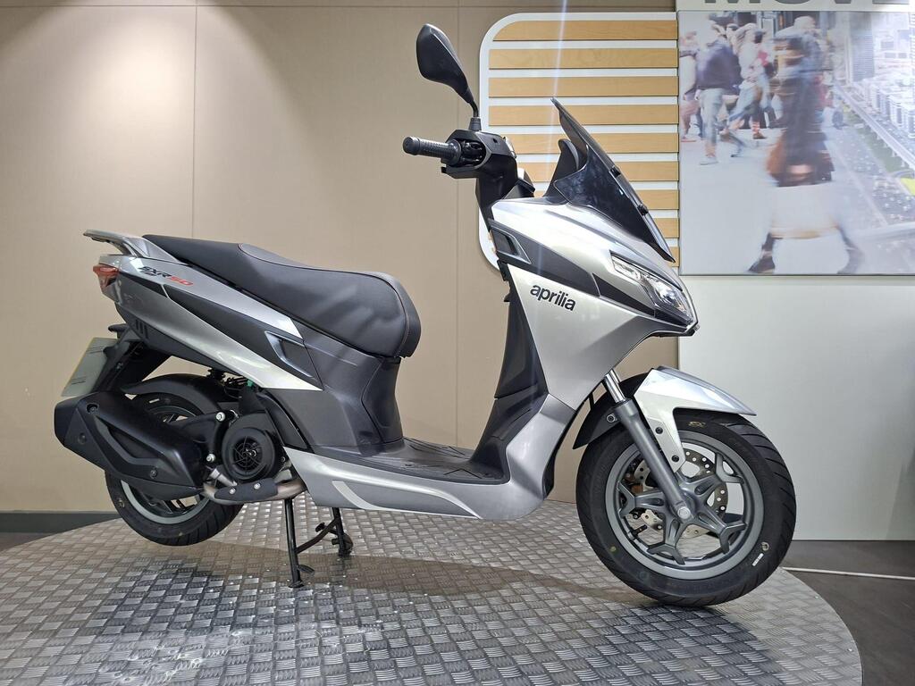 Aprilia SXR 50 2026 5 A silver Aprilia scooter displayed on a circular metal platform, with a minimalist background featuring a blurred image of people.