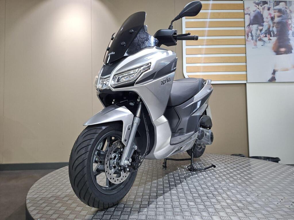 Aprilia SXR 50 2026 6 A silver Aprilia scooter displayed on a rotating platform in a modern showroom. The scooter features a sleek design with a transparent windscreen, stylish headlight, and a wide front tire. Background elements include a plain wall and a partially visible image of people in a busy urban setting.