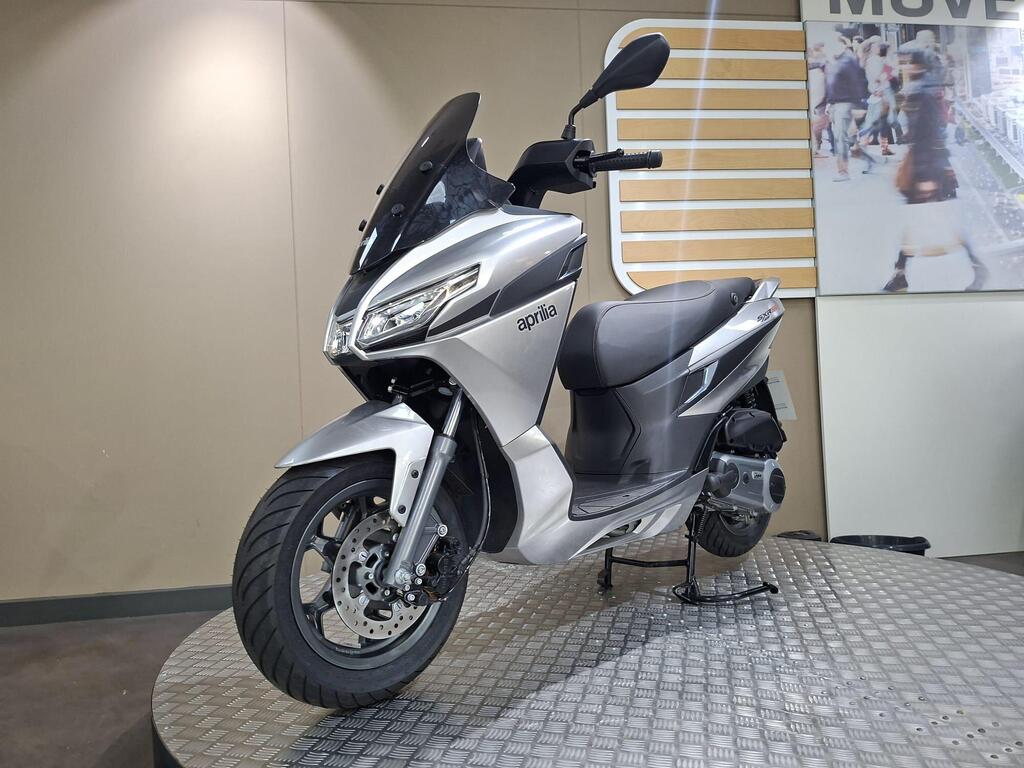 Aprilia SXR 50 2026 7 A silver Aprilia scooter displayed on a circular platform, set against a neutral background with a partially visible image of people in motion on the wall behind.